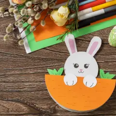 Uroczy króliczek z papieru kolorowego wychyla się z marchewki. Obok pisanki z papieru kolorowego, bazie i flamastry.