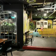 Warszawska restauracja z salą zabaw dla dzieci. Widać stoliki, krzesła i kolorowy plac zabaw z domkiem i zjeżdżalnią.