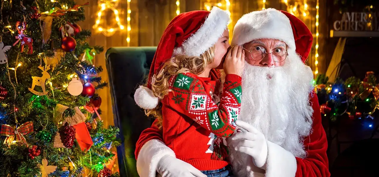 Mała dziewczynka szepcze życzenia do ucha Świętego Mikołaja, siedząc przy choince. To magiczny moment dla dzieci.