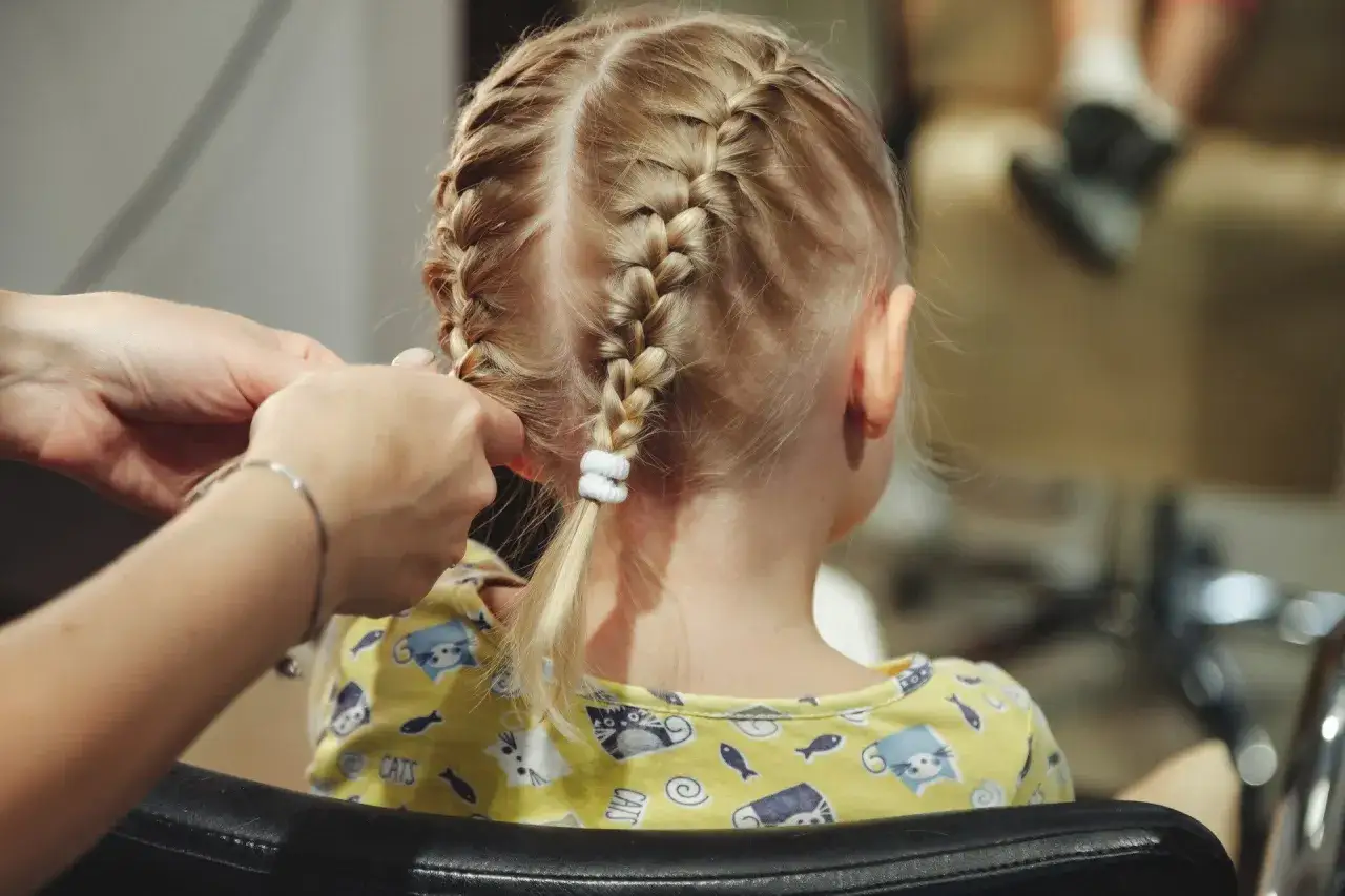 Ręce zaplatają dwa warkocze na blond włosach dziewczynki. To świetny sposób, jak uczesać dziewczynkę na co dzień.