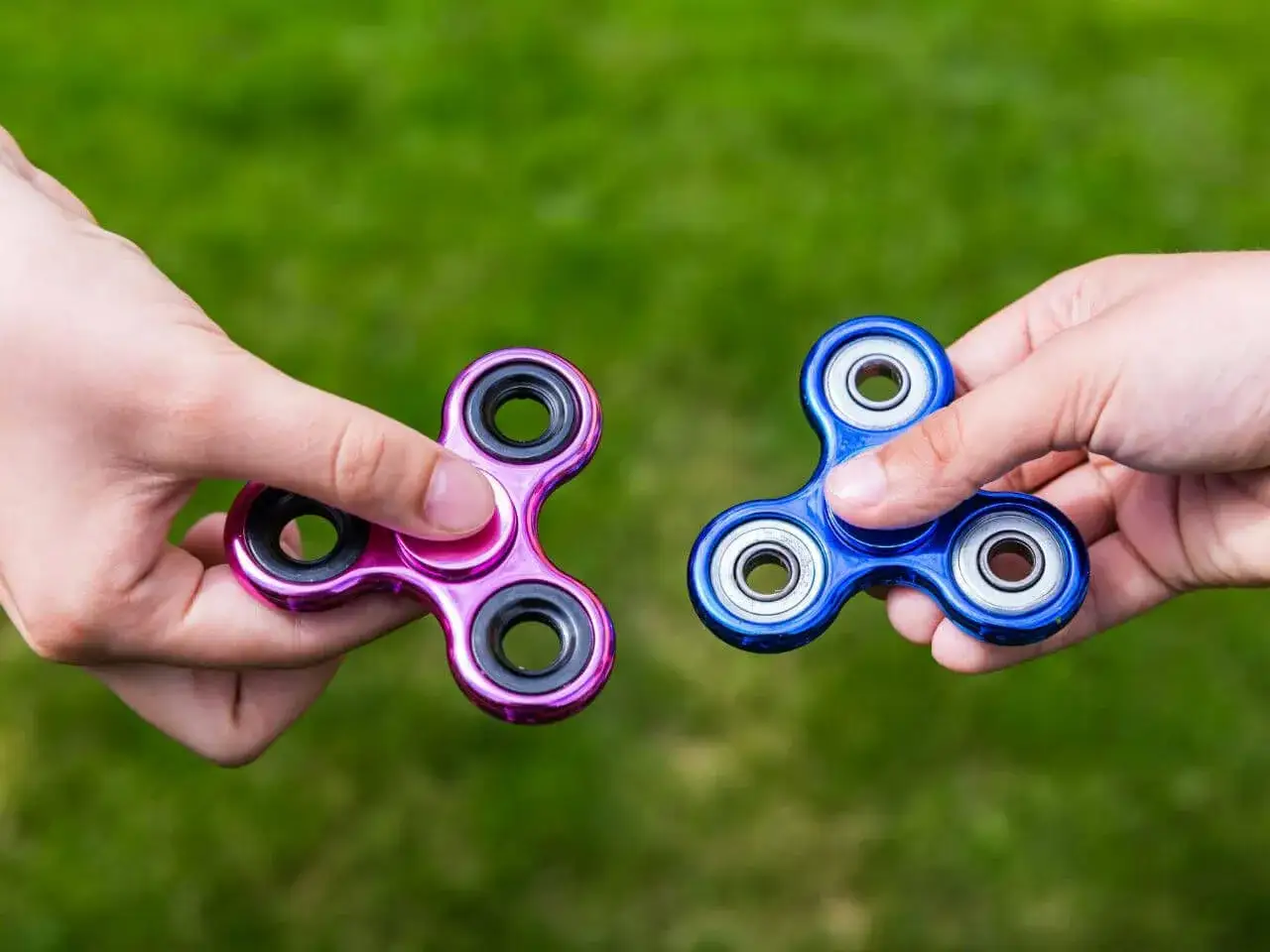 Dwie ręce trzymają kolorowe spinner dla dzieci na tle zielonej trawy.